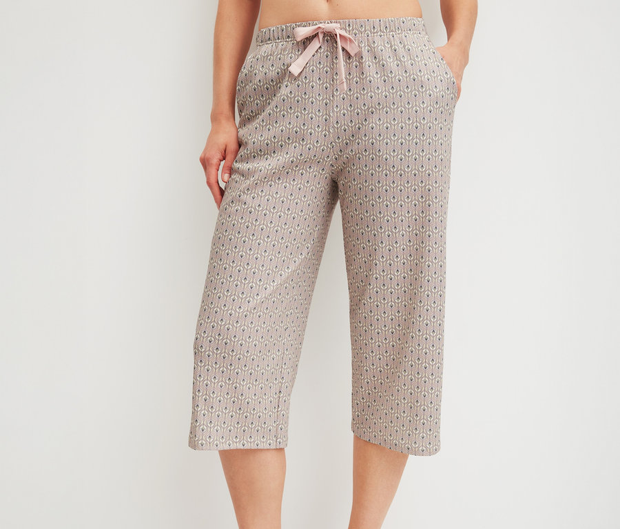 Eine Person trägt eine gemusterte CALIDA »Favorites Arts« 3/4-Hose in Beige mit rosa Kordelzug und hat eine Hand in der Tasche. Die nackten Füße sind sichtbar.