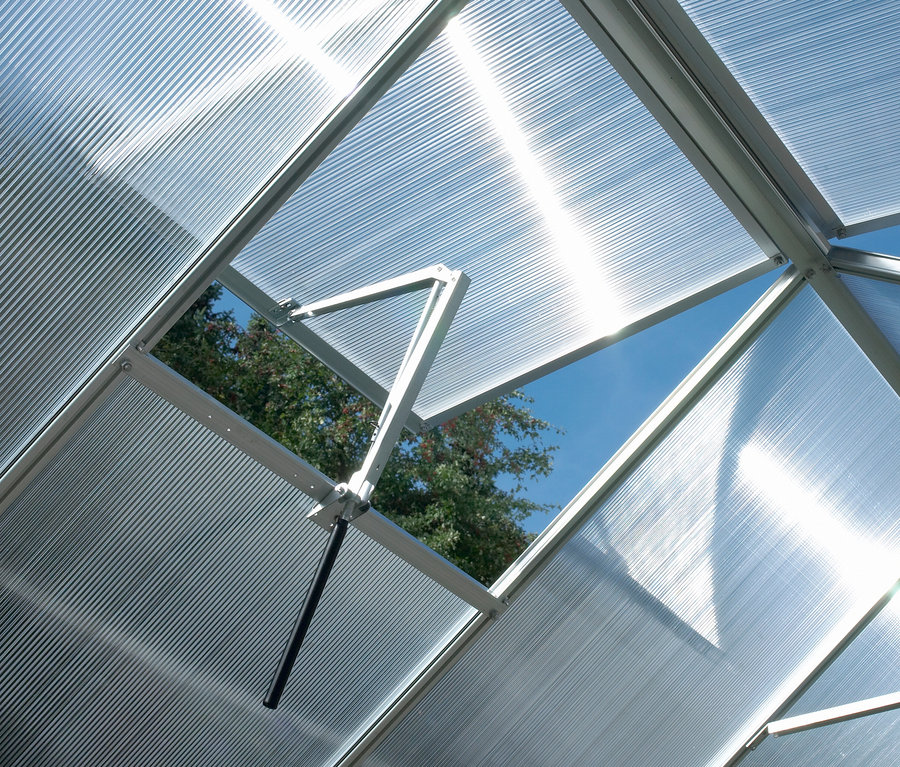 Innenansicht eines Gewächshauses mit geöffnetem Vitavia automatischen Fensterheber »Thermovent«, durch den Himmel und Baumwipfel sichtbar sind.