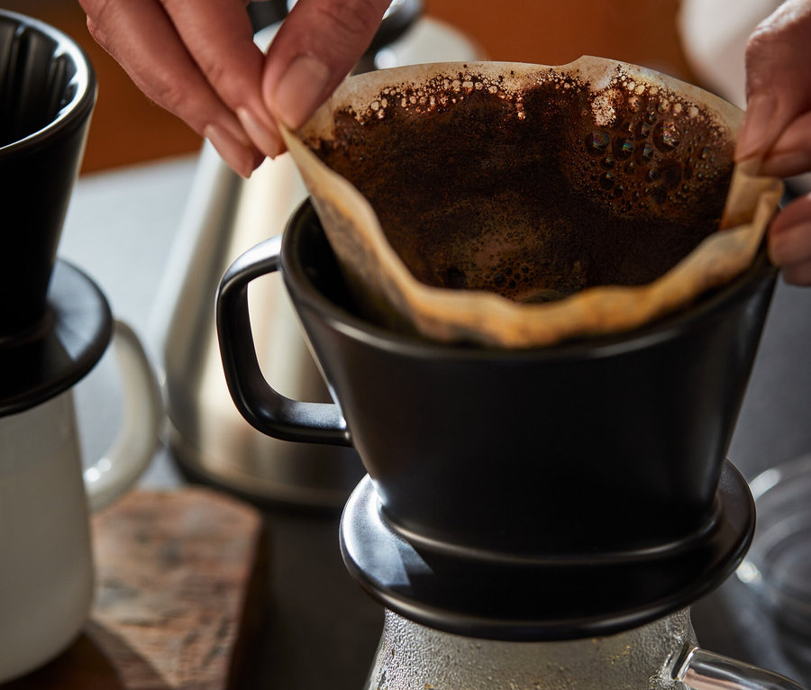 Frau legt Papierfilter in schwarzen Kaffee Handfilter Gr. 2. Darunter steht eine Kaffeekanne aus Glas.