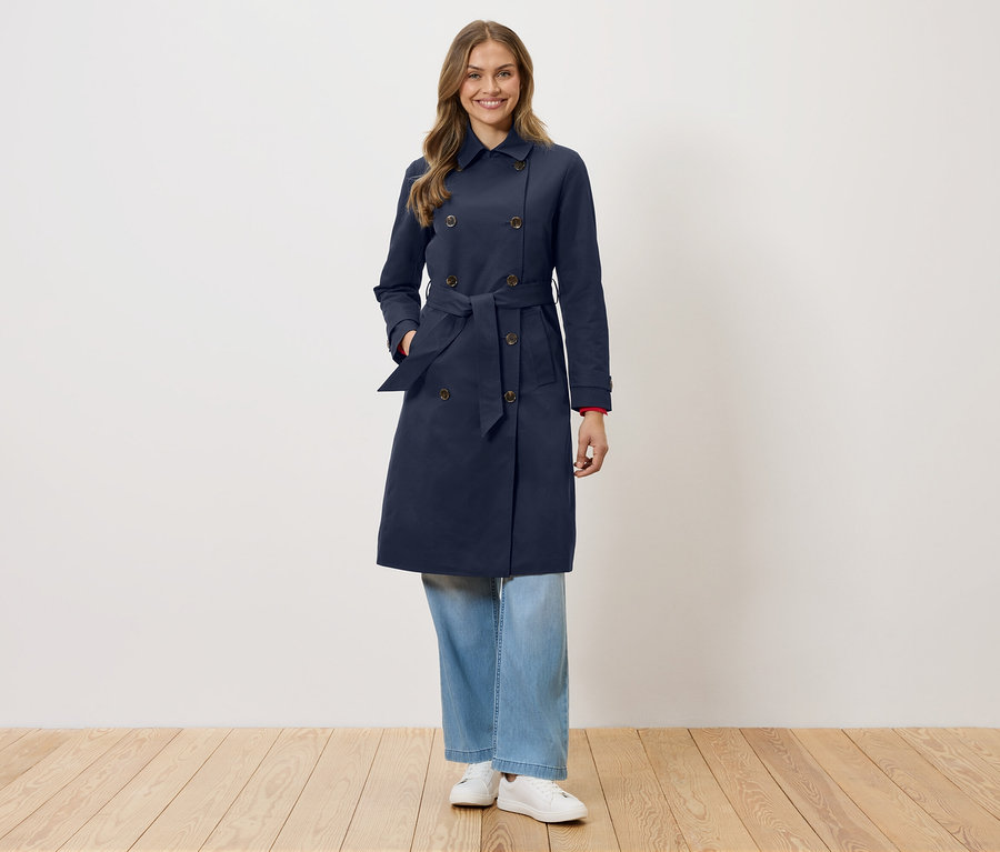 Eine Frau steht vor einer weißen Wand und trägt einen marineblauen Trenchcoat mit Gürtel, blaue Jeans und weiße Turnschuhe.
