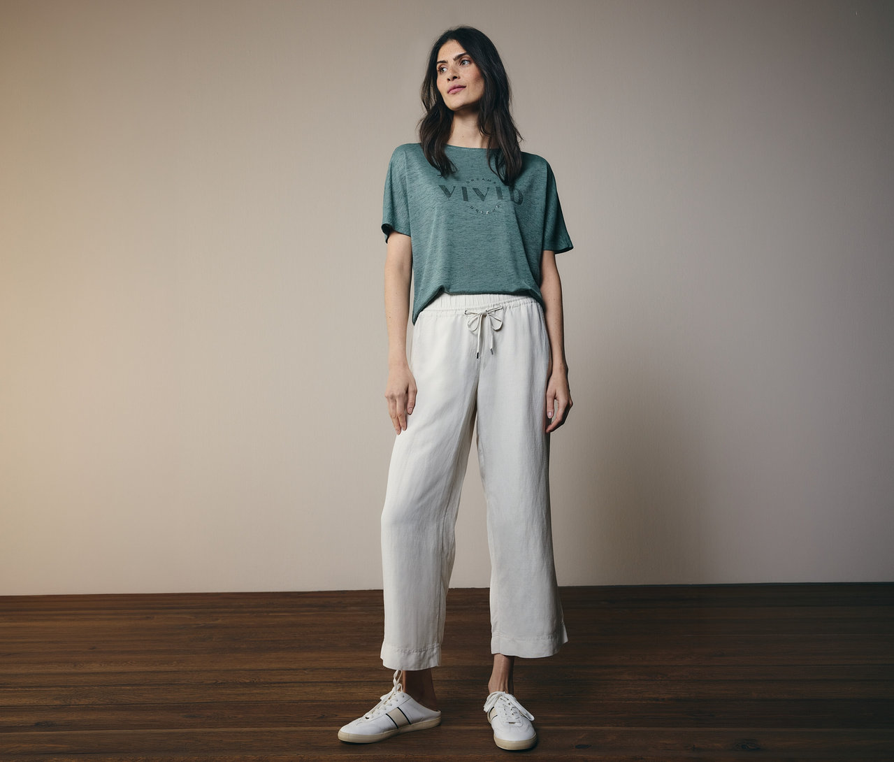 Eine Frau steht in einem grünen T-Shirt mit „VIVID“-Aufdruck, beigen weiten Hosen mit Kordelzug und weißen Slip-on-Sneakern.