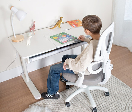 Junge sitzt an einem weißen Schreibtisch auf einem weißen Bürostuhl. Auf dem Tisch liegen ein Buch, eine Lampe und Spielzeugtiere.