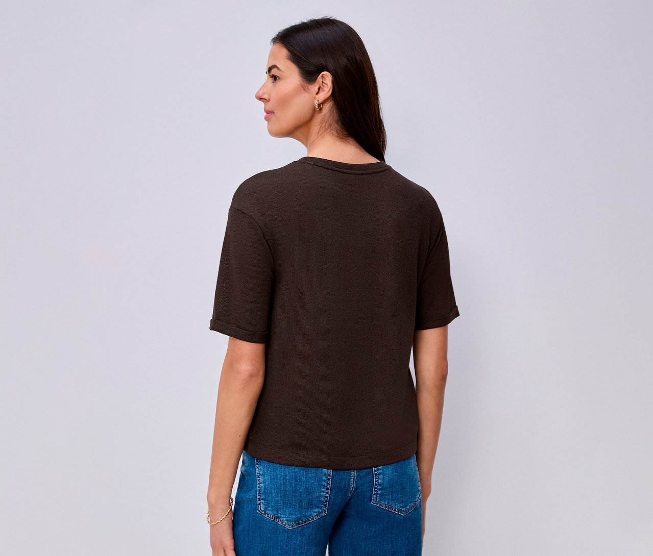 Eine Frau von hinten, leicht nach rechts gedreht, trägt ein dunkelbraunes Street One T-Shirt mit kurzen Ärmeln und blaue Jeans. Sie hat lange dunkle Haare, kleine goldene Ohrringe und ein goldenes Armband am linken Handgelenk.