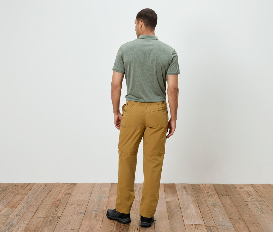 Ein Mann steht mit dem Rücken zur Kamera, er trägt ein olivgrünes Poloshirt und eine goldene Outdoorhose mit Reißverschluss. Er steht auf einem Holzboden.