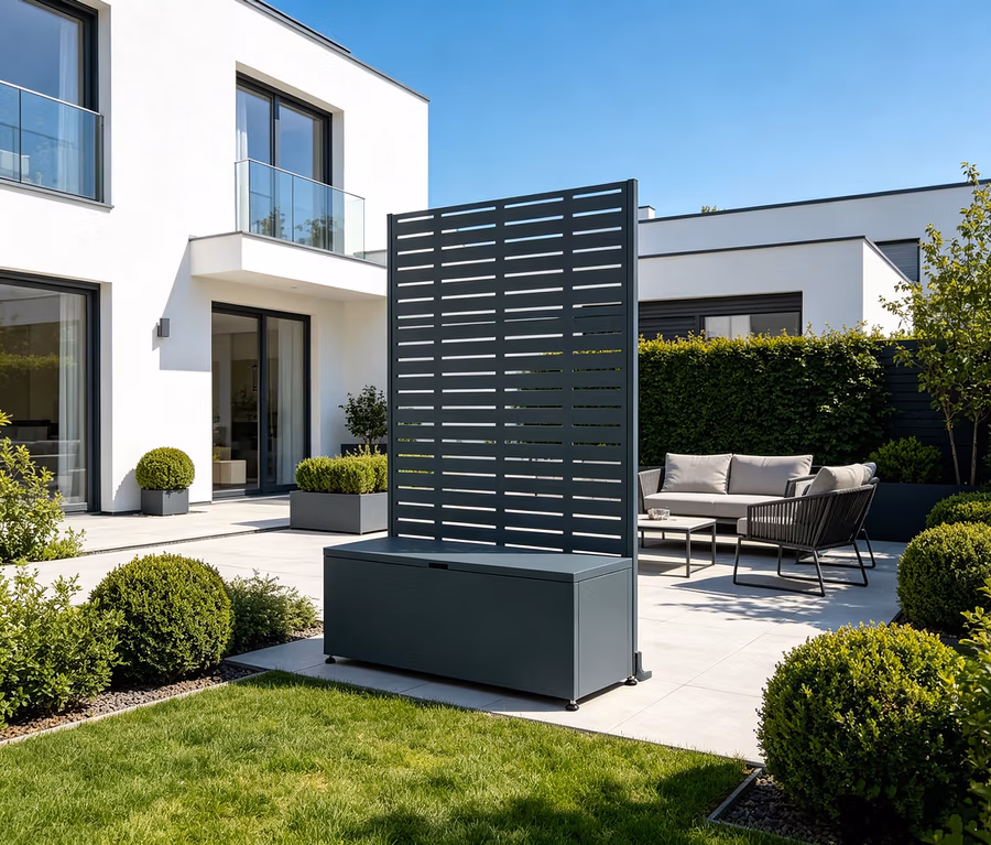 Ein moderner Außenbereich mit einem dunkelgrauen Sichtschutz mit horizontalen Lamellen, der in eine darunterliegende Aufbewahrungsbox integriert ist. Der Sichtschutz steht auf einer hellen Terrasse neben einem grünen Rasen. Im Hintergrund sind ein weißes, modernes Haus mit großen Fenstern und Balkon sowie eine Sitzgruppe mit Sofa, Sessel und Couchtisch zu sehen.