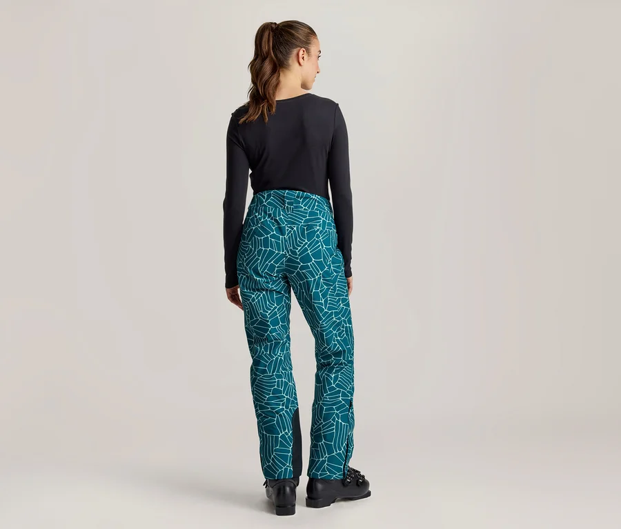 Frau von hinten stehend, trägt ein schwarzes Langarmshirt und eine petrolfarbene Hose mit weißem geometrischem Muster sowie schwarze Stiefel.