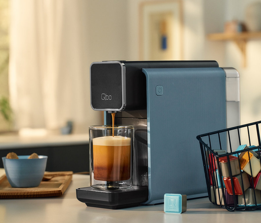 Kaffee läuft aus einer Qbo-Kaffeemaschine in ein Glas. Eine Schale mit Zuckerwürfeln und ein Kapselkorb stehen auf der Theke.