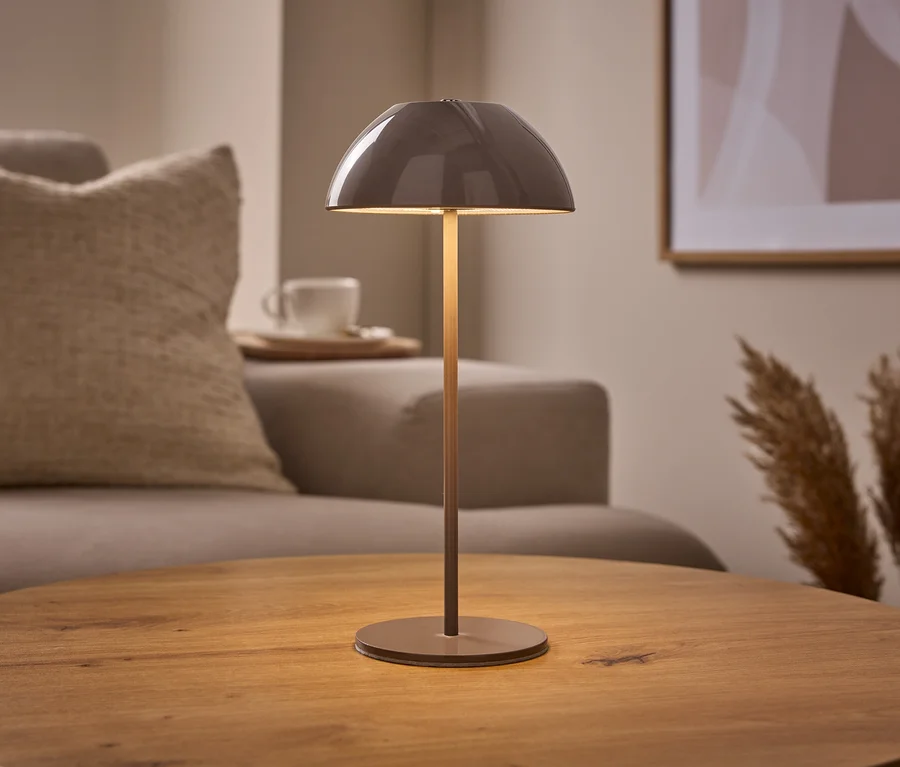 Eine beige LED-Akku-Tischleuchte mit gewölbtem Schirm steht auf einem runden Holztisch. Im Hintergrund sind ein Sofa, eine Tasse und Pampasgras zu sehen.