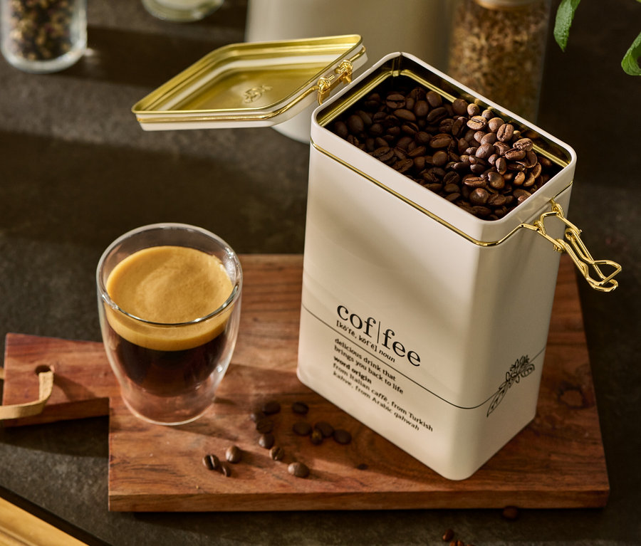 Eine geöffnete Kaffeedose, coffee dictionary, gefüllt mit Kaffeebohnen, steht neben einem Glas Kaffee auf einem Holzbrett.