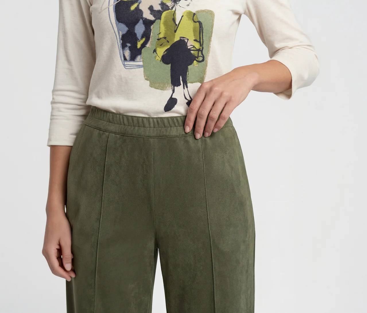 Nahaufnahme einer Person, die ein cremefarbenes Langarmshirt mit Grafikdruck und olivgrüne Hose trägt. Die Hand der Person ruht auf dem elastischen Bund der Hose, die vertikale Nähte hat.