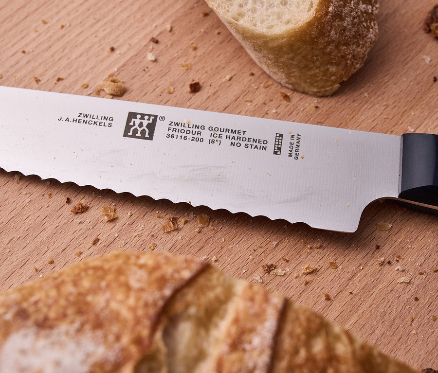 Ein gezacktes Brotmesser von ZWILLING GOURMET liegt diagonal auf einem Holzschneidebrett, umgeben von Brotkrümeln und zwei Stücken Krustenbrot.