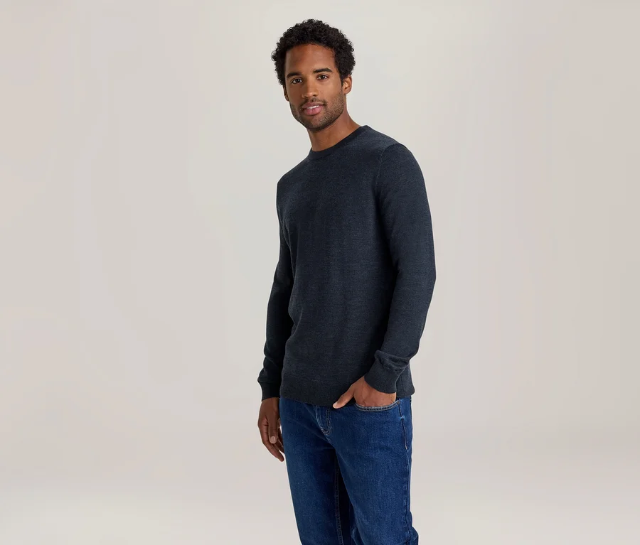 Mann in dunkelblauem Pullover und Jeans, eine Hand in der Hosentasche.