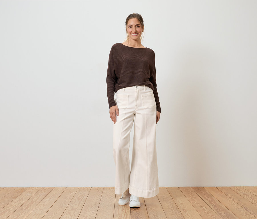 Eine lächelnde Frau trägt einen braunen Pullover und eine weiße Wide Leg Jeans im Fit „Viki“.