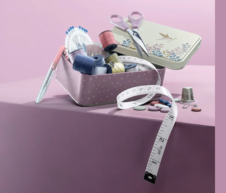 Näh-Set in Metallbox mit Schere, Knöpfen, Nadeln und Maßband auf lila Hintergrund.