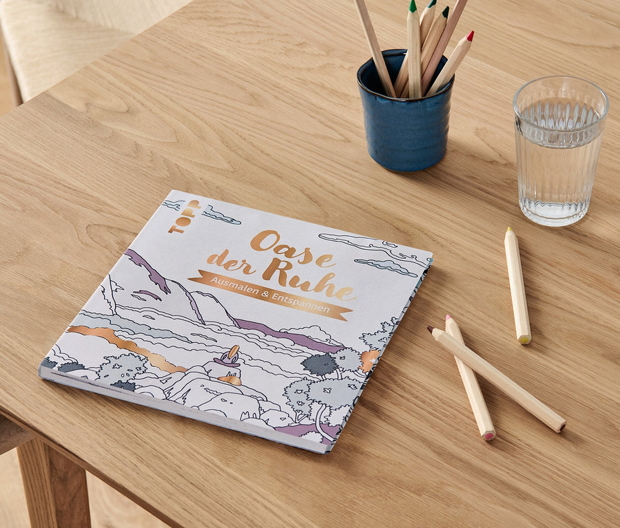 Auf einem Holztisch liegen das Buch "Oase der Ruhe - ausmalen und entspannen", ein Glas Wasser und ein Becher mit Buntstiften.
