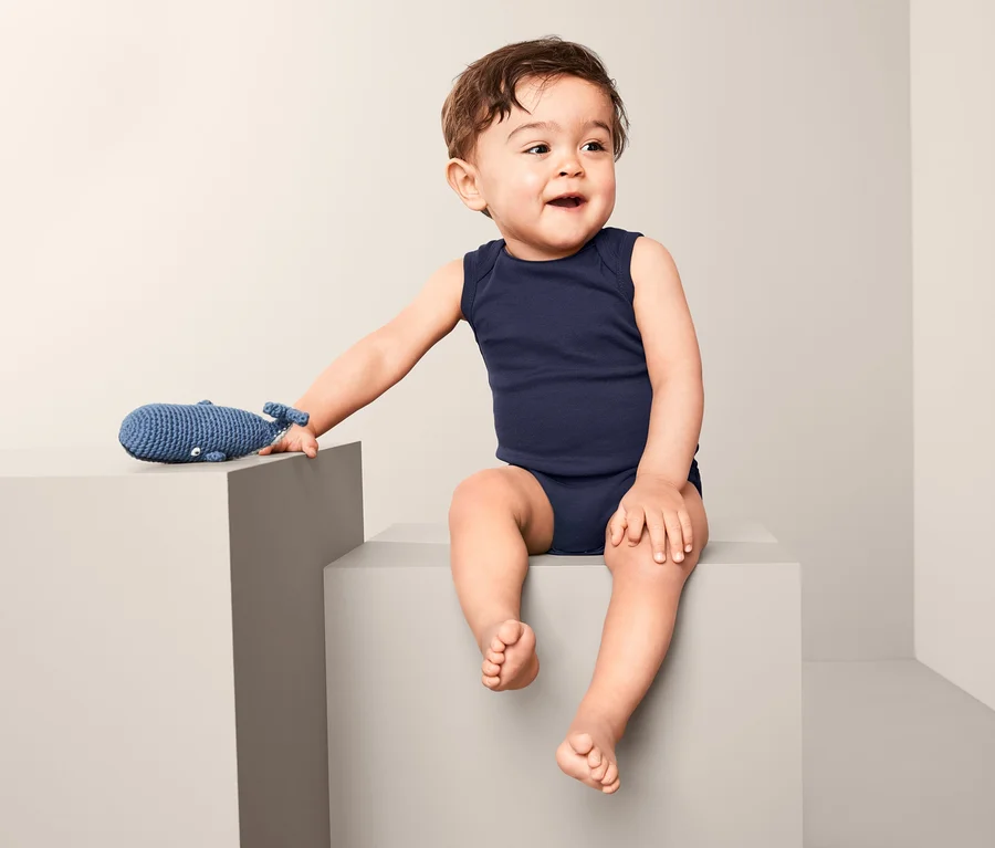 Ein Baby sitzt auf einem grauen Würfel und trägt einen blauen, ärmellosen Body. Neben ihm liegt ein blaues Wal-Spielzeug.