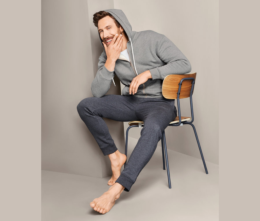 Mann sitzt barfuß auf einem Stuhl, lacht und trägt eine Kapuzen-Sweatjacke.