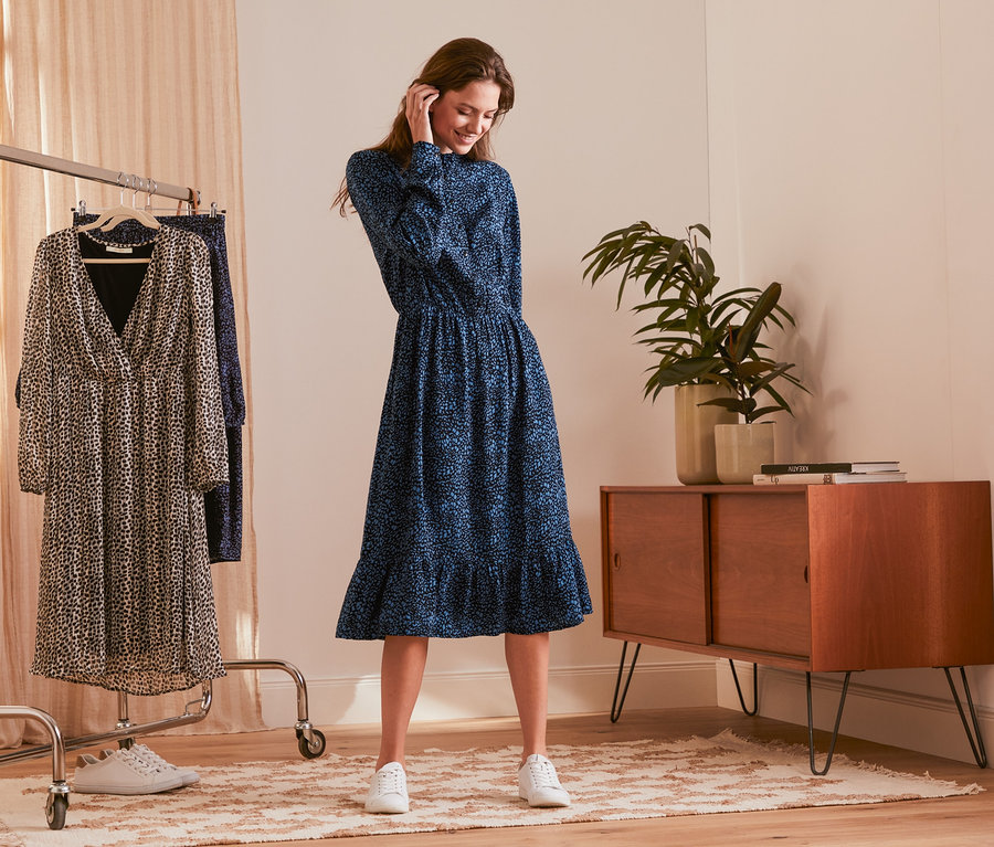 Frau trägt ein blaues Hemdblusenkleid. Zwei Kleider hängen auf einem Kleiderständer.