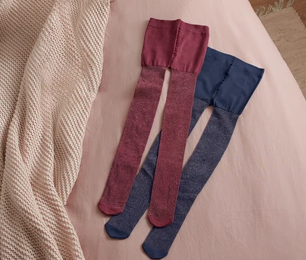 Zwei Paar Kleinkinder-Strumpfhosen liegen auf einem rosafarbenen Bettlaken neben einer beigen Strickdecke.