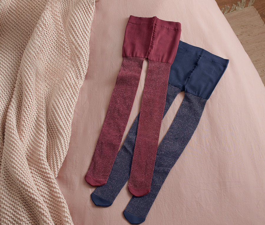 Zwei Paar Kleinkinder-Strumpfhosen liegen auf einem rosafarbenen Bettlaken neben einer beigen Strickdecke.