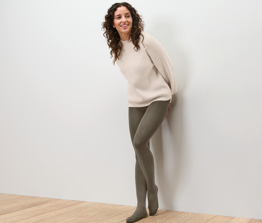 Frau lehnt an Wand und trägt Thermostrumpfhose und Pullover.