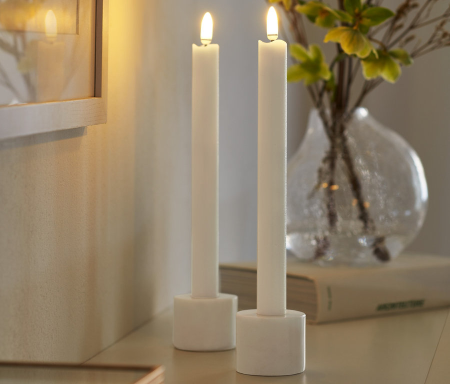 Zwei LED-Echtwachs-Stabkerzen stehen auf einem Tisch neben einer Vase mit Blumen.