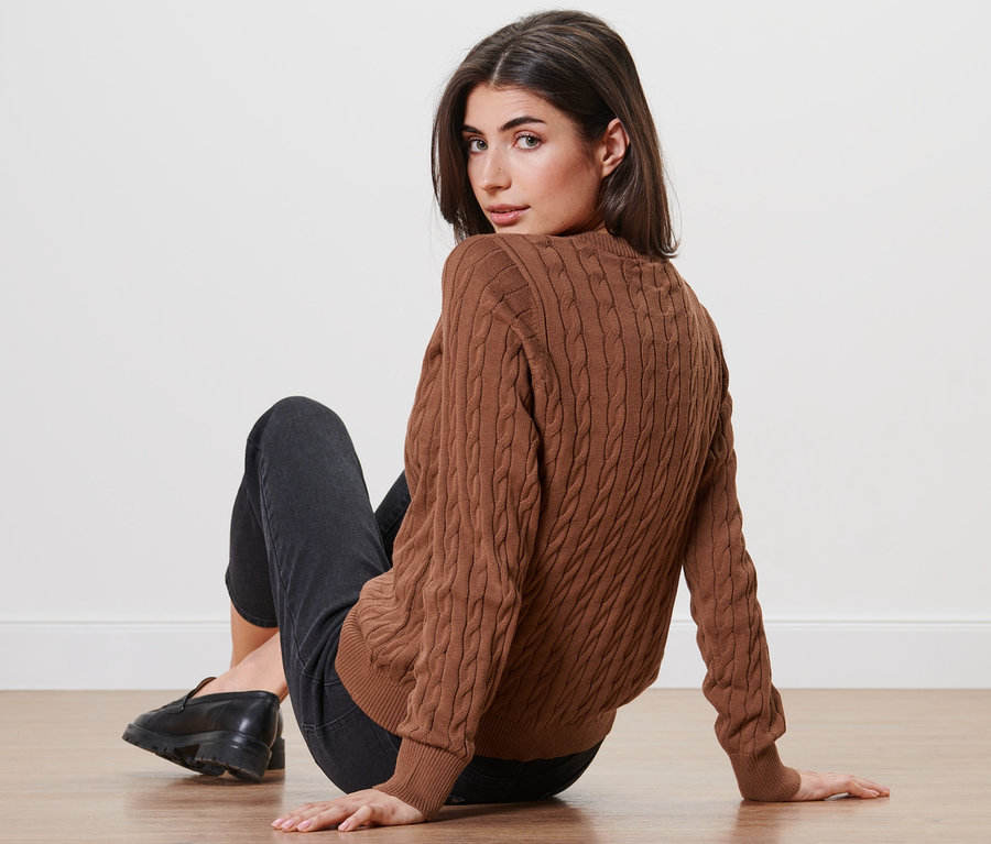 Frau sitzt auf dem Boden und blickt über die Schulter, trägt einen Feinstrickcardigan mit Zopfmuster.