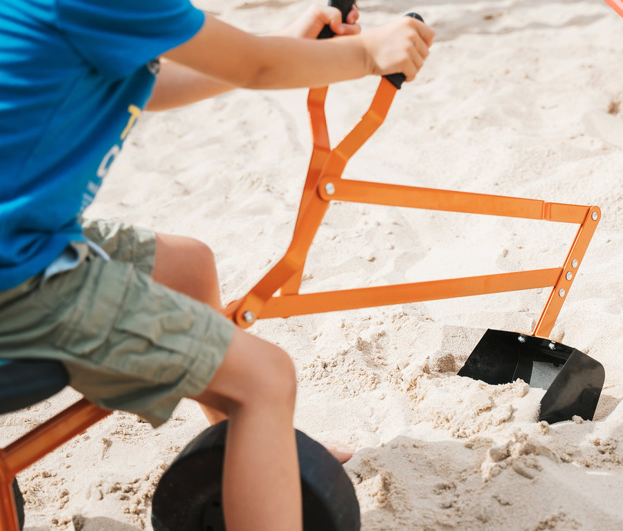 Junge sitzt auf einem orangefarbenen Small Foot Bagger mit Rädern und spielt im Sand.