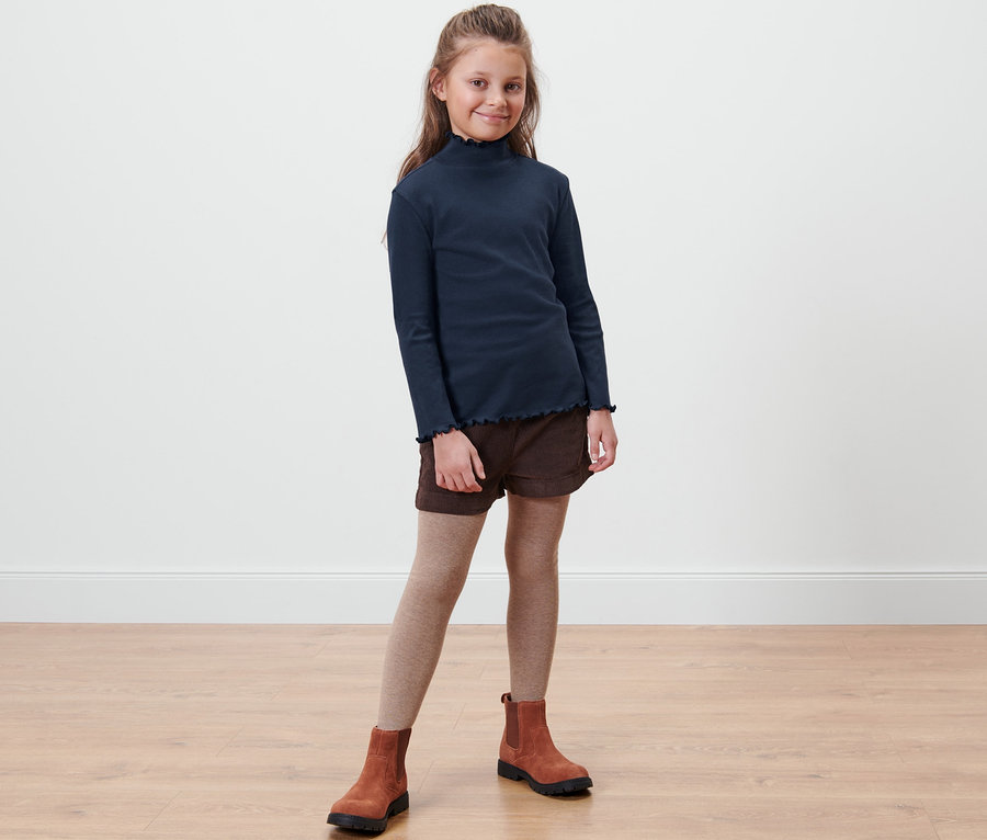 Mädchen steht und trägt ein Kinder-Langarmshirt, braune Shorts, braune Stiefel und beige Strumpfhosen.