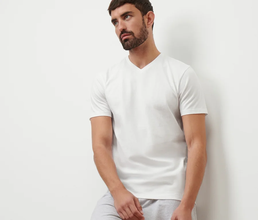 Mann lehnt an einer Wand, trägt graue Shorts und ein weißes T-Shirt mit V-Ausschnitt.