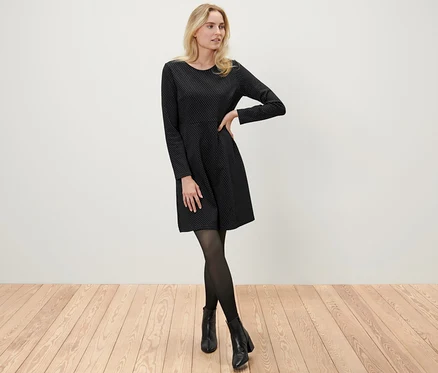 Blonde Frau posiert in einem glitzernden Jacquard-Kleid, schwarzen Strumpfhosen und Stiefeletten.