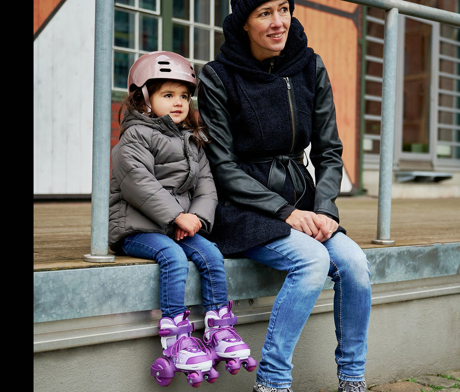 Mutter und Tochter sitzen nebeneinander. Das Mädchen trägt einen rosa Helm und lila HUDORA Rollschuhe »My First Quad«.