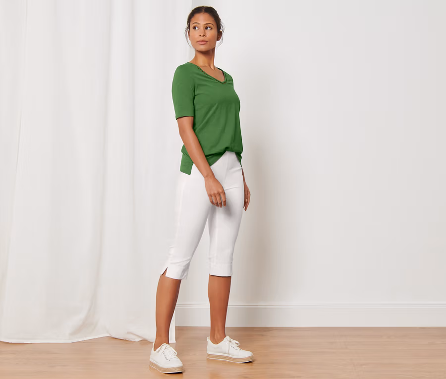 Eine Frau steht in einem Raum und trägt ein grünes T-Shirt und eine weiße 3/4-Stretchhose.