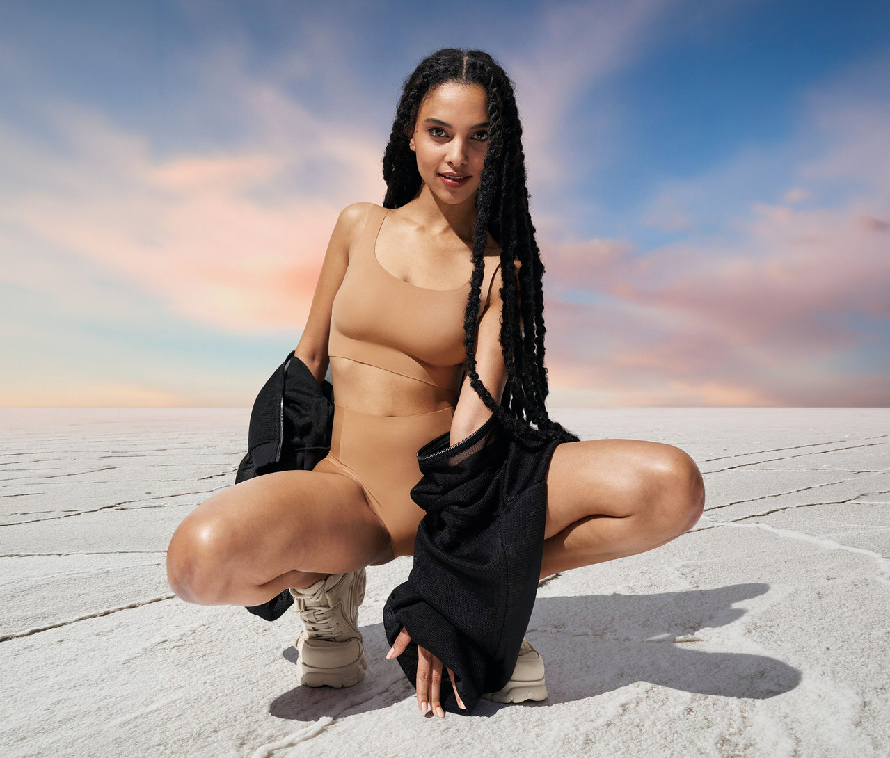 Frau hockt in braunem Sloggi ZERO Feel 2.0 Top vor blau-rosa Himmel. Sie hält eine schwarze Jacke.