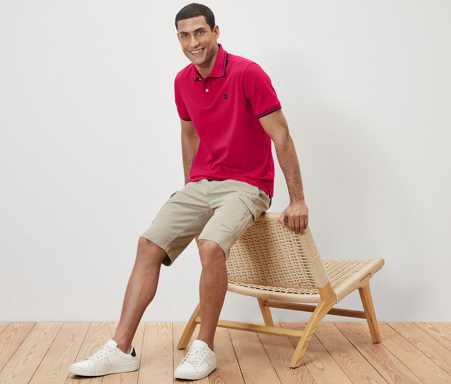 Mann sitzt auf einem Korbstuhl und trägt ein rosa Piqué-Poloshirt.