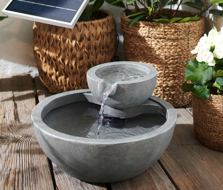 Ein Solar-Gartenbrunnen steht auf einem Holzboden, im Hintergrund Pflanzen in geflochtenen Körben.