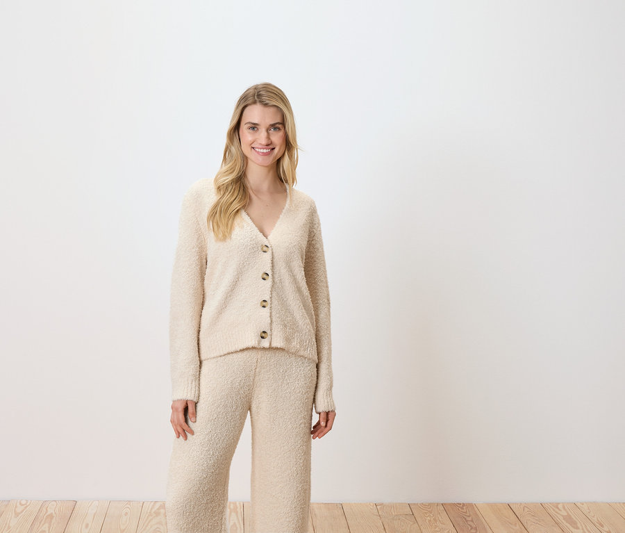 Frau posiert vor einem weißen Hintergrund und trägt eine cremefarbene Strickjacke und Hose.