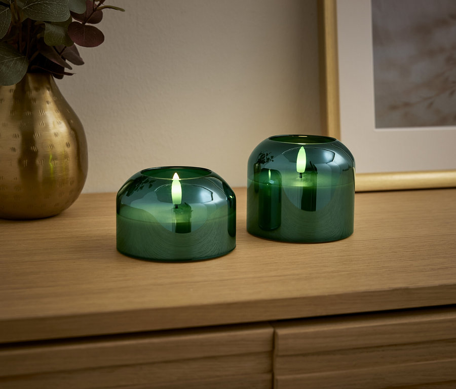 Zwei grüne Kerzen und eine goldene Vase stehen auf einem Holztisch.