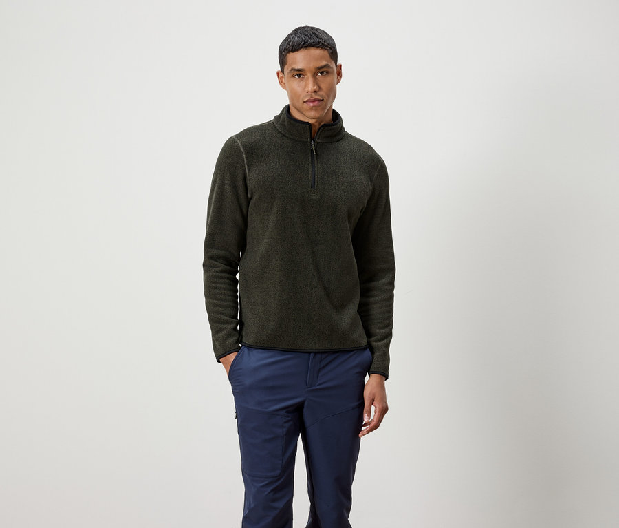 Mann steht und trägt einen olivgrünen Pullover mit Reißverschluss und blaue Hosen.