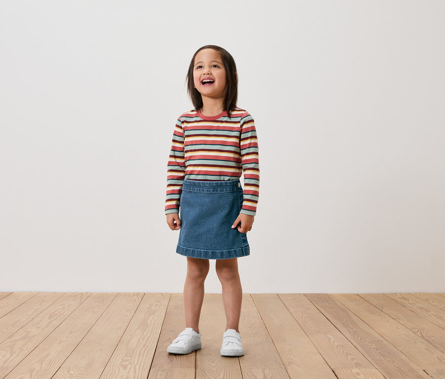 Ein Mädchen steht auf einem Holzboden und trägt ein langärmliges Ringelshirt, einen Jeansrock und weiße Turnschuhe.