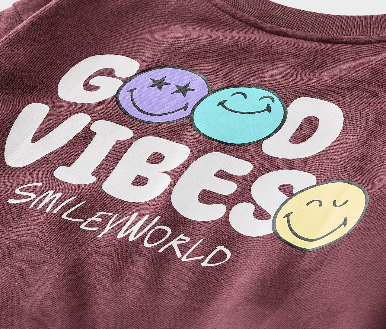 Detail eines bordeauxroten Kinder-Sweatshirts SMILEYWORLD® mit angerauter Innenseite und dem Aufdruck GOOD VIBES und drei Smileys.