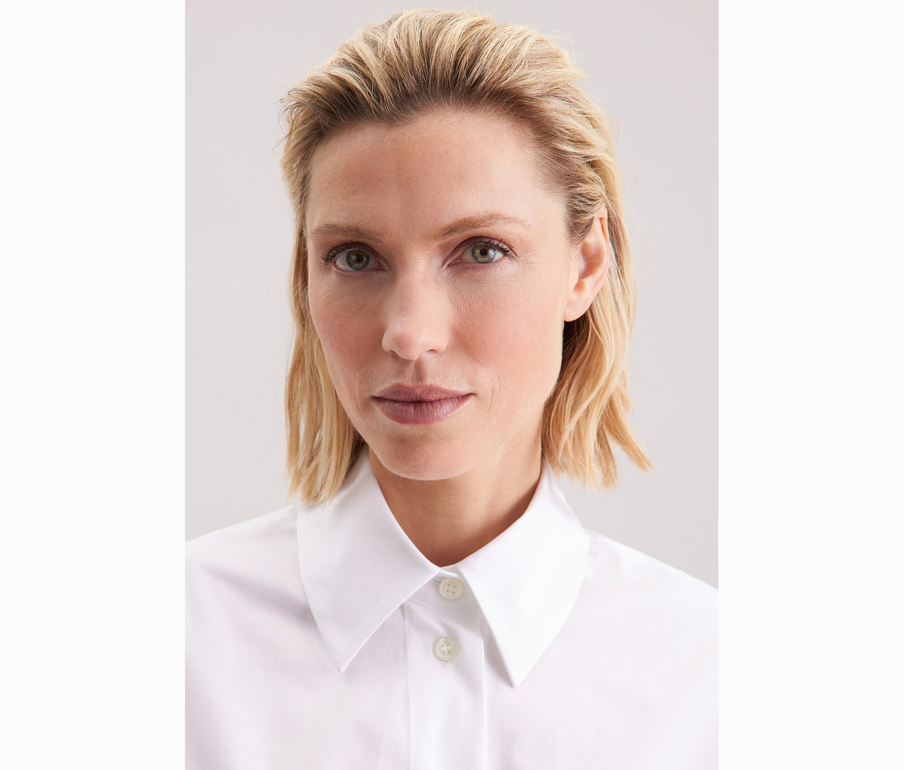 Porträt einer Frau mit blonden, gewellten Haaren, die eine weiße Seidensticker Oversize Longbluse trägt.