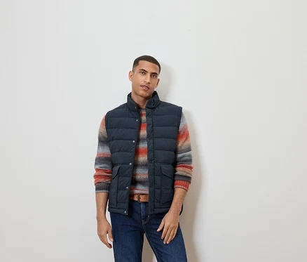 Mann trägt eine Steppweste, einen gestreiften Pullover und Jeans.