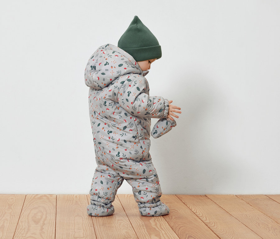 Kleinkind in grauem Overall mit Blumenmuster und grüner Mütze.