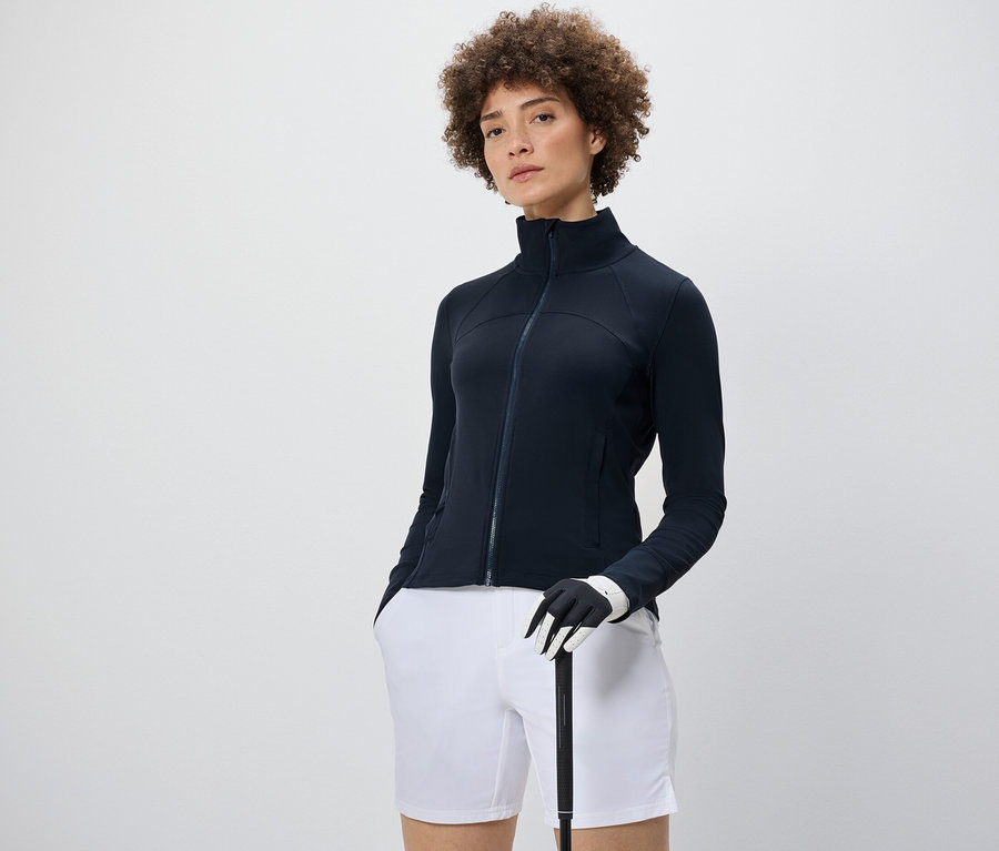 Frau trägt eine Funktionsjacke, weiße Shorts und hält einen Golfschläger.