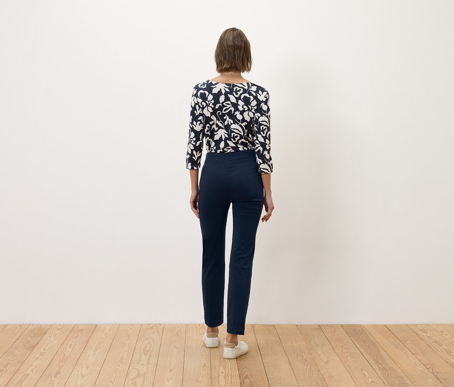Eine Frau mit braunen Haaren steht mit dem Rücken zur Kamera und trägt eine marineblaue Hose und ein blau-weißes Oberteil mit Blumenmuster.