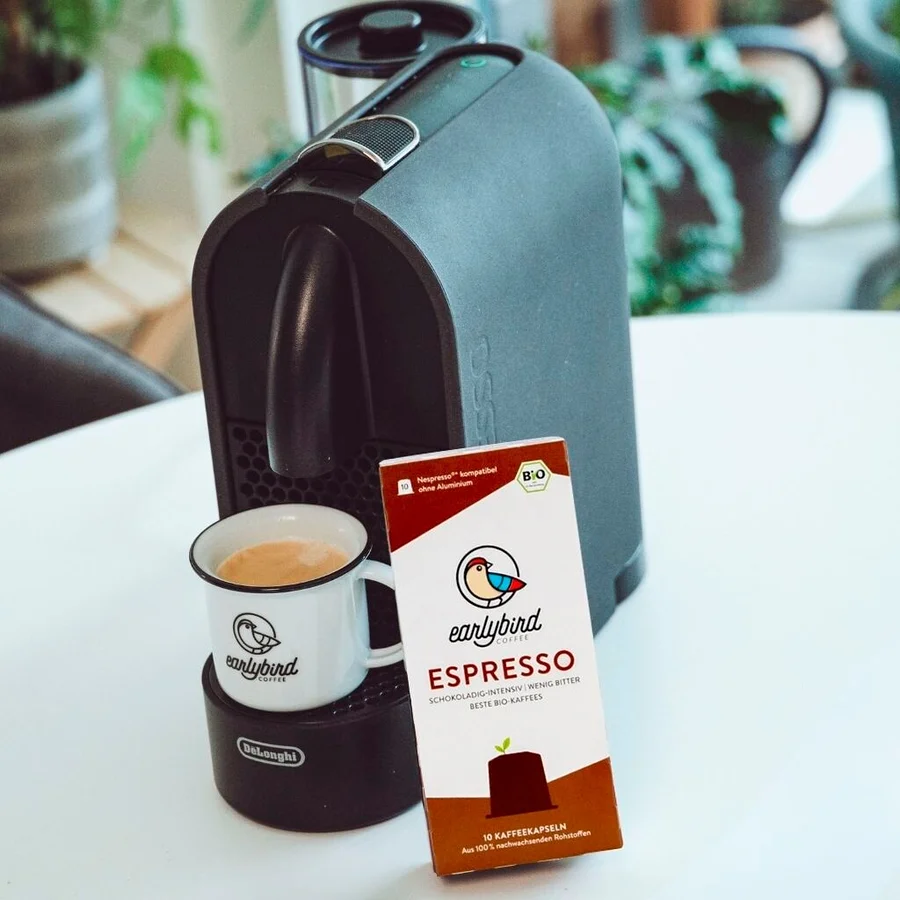 Eine Tasse Kaffee steht auf einer DeLonghi Maschine, daneben eine Packung earlybird Bio Espresso Kapseln.