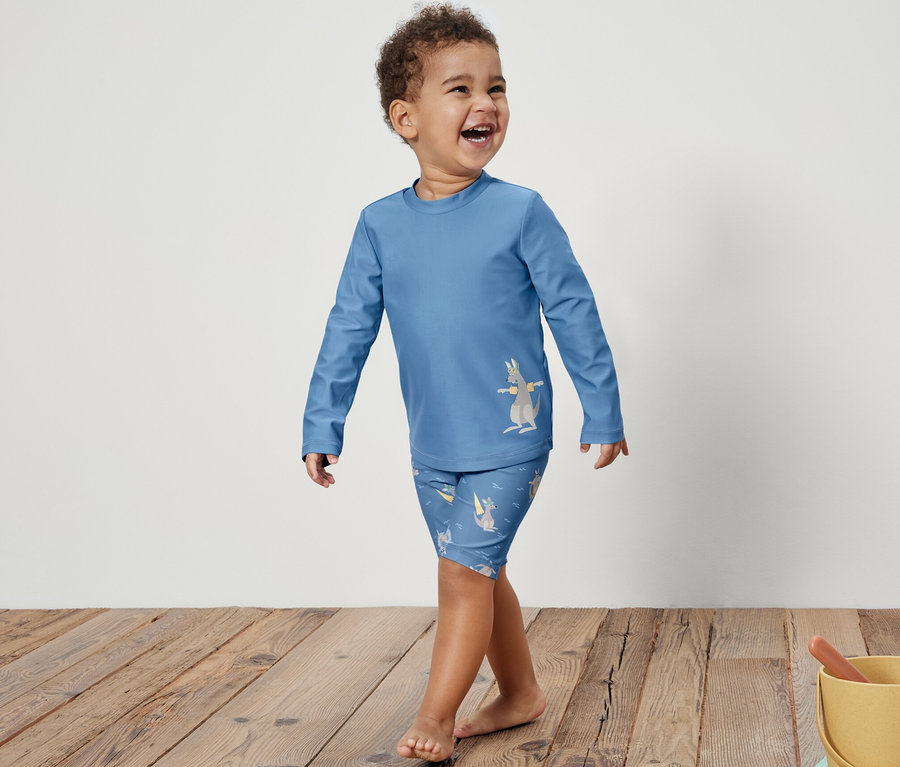 Ein lächelnder kleiner Junge in einer blauen Kinder-Kombi mit UV-Schutzfaktor 80 und Känguru-Print geht auf einem Holzboden. Rechts steht ein gelber Eimer mit Spielzeug im Sand.
