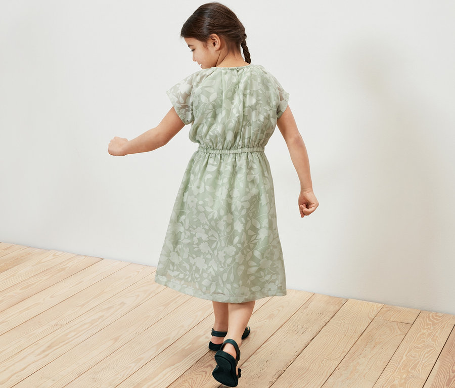 Mädchen geht in einem grünen Kinder-Kleid über einen Holzboden.
