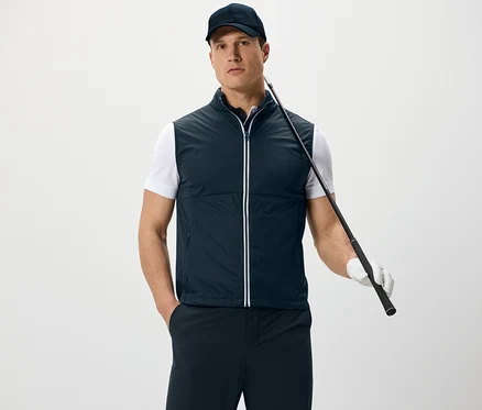 Ein Mann trägt eine Softshellweste und eine Kappe, hält einen Golfschläger und steht vor einem weißen Hintergrund.